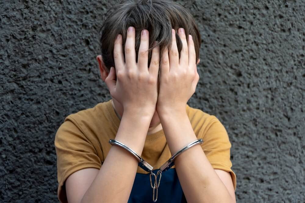 young boy in handcuffs sits on a gray background. Juvenile delinquent, criminal liability of minors.