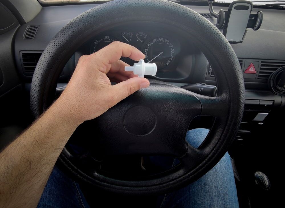 man's hand holding a breathalyzer piece in front of a car steering wheel